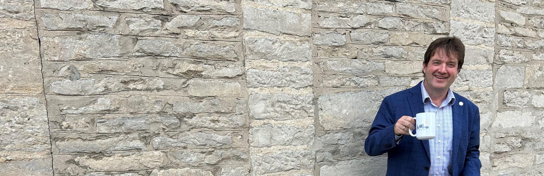 Mayor standing in front of stone wall with a cup of coffee